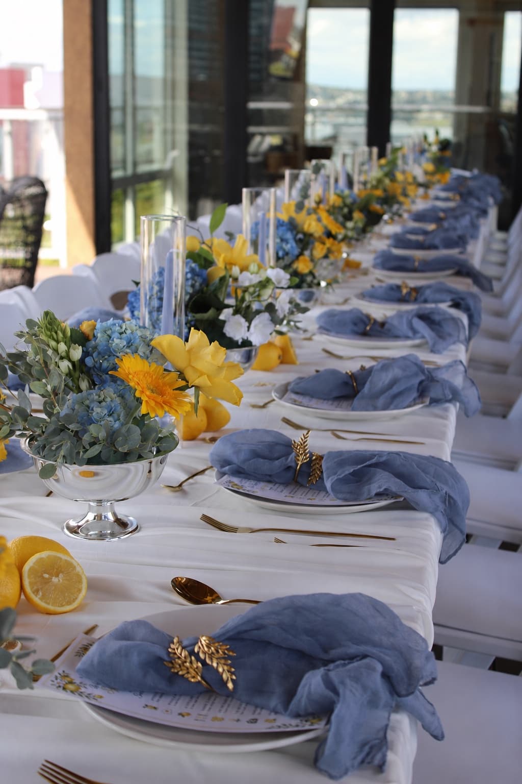 An elevated waterfront banquet table styled by Events by Juna with white linens, dusty-blue gauze napkins in gold napkin rings, blue hydrangea and yellow rose centerpieces in footed silver bowls, and gold flatware.